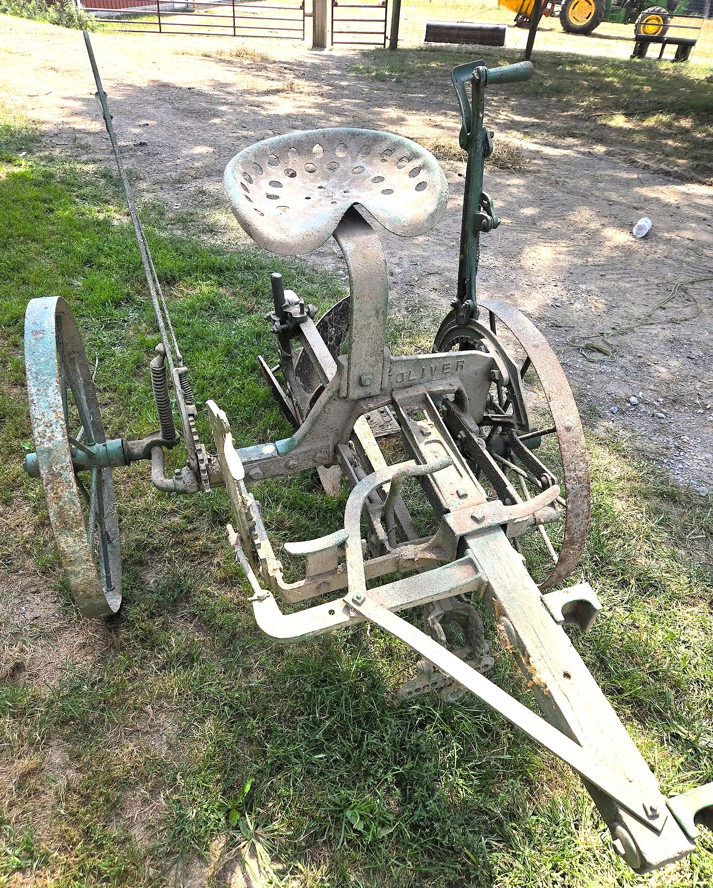 Old rusty pedal-powered farm implement with a rounded perforated saddle and crank-driven wheels in a grassy yard at a farm.